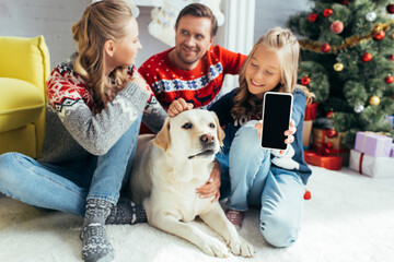joyful kid cuddling dog while holding smartphone with blank screen near parents in decorated living room