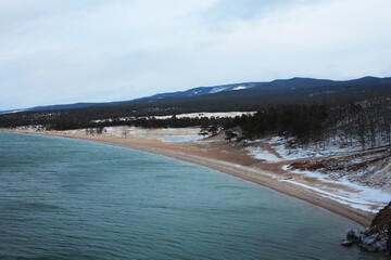 The beach is late autumn, in November. Snow lies on the sand, the water of Lake Baikal is turquoise, with mountains in the backgroun