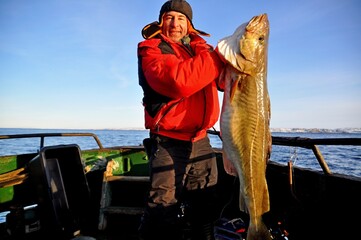 A happy fisherman in a red jacket caught and holds in his hands a beautiful large huge cod on a...