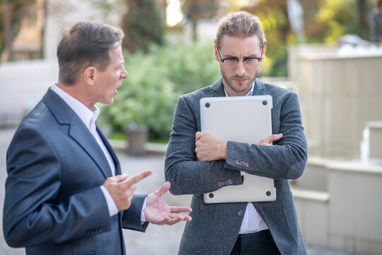 Angry Male Arguing With His Younger Male Colleague Holding Laptop Outside