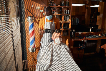 Barber in medical mask cutting client hair in barbershop
