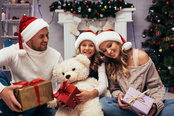 Obraz na płótnie Canvas happy kid holding teddy bear near presents and parents in santa hats