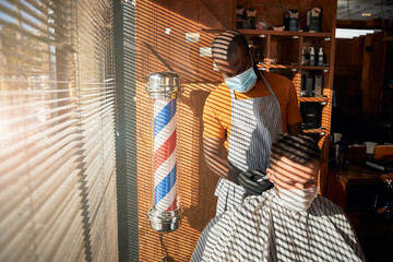 Barber in medical mask making haircut for man in barbershop