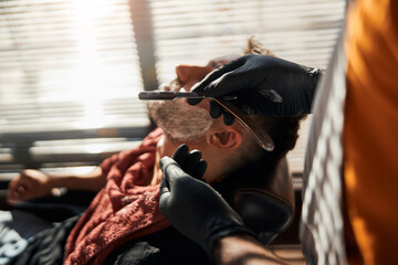 Barber in sterile gloves shaving man beard with straight razor