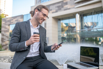 Male office worker sitting with coffee cup near fountain, checking his phone