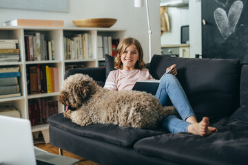 teenage girl sitting on sofa at home and reading book with her dog