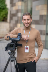Smiling fair-haired cameraman standing next to camera, leaning on elbow