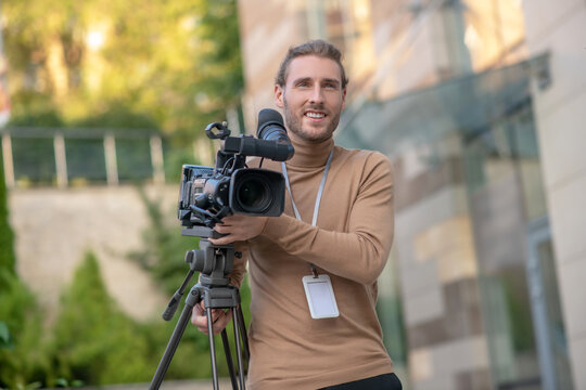 Smiling fair-haired cameraman carring camera with tripod outside