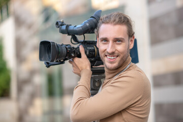 Smiling fair-haired operator standing with camera on his shoulder outside