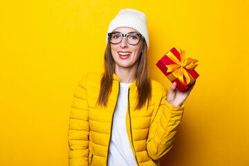 Smiling young woman in yellow jacket holding gift on yellow background.