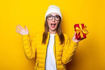Surprised shocked young woman in yellow jacket holding gift on yellow background.