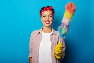 Smiling young woman in gloves holding dust brush on blue background.