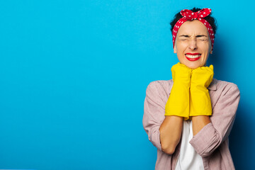 Young woman in a red bandage for hair, in gloves for cleaning screwing up is eyes on a blue background.