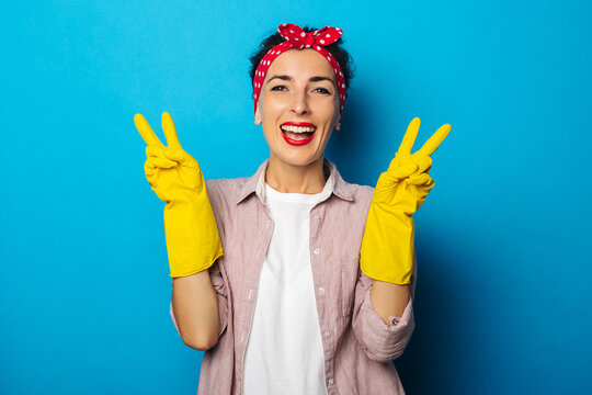 Smiling young woman in cleaning gloves shows two fingers gesture, peace gesture, victory on blue background.