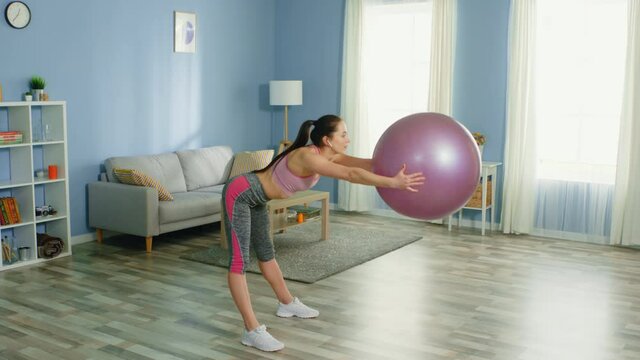 Pretty woman is doing exercise with gym ball, practicing pilates at home, listening her favorite music, in positive mood, enjoying her active morning, slow motion.