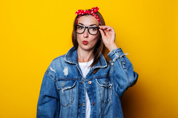 Young woman in hair band, in a denim jacket holding glasses making lips with a bow on a yellow background.