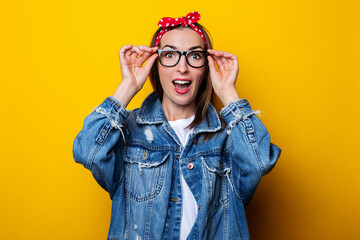 Surprised young woman in hair band, in denim jacket holding glasses on yellow background.