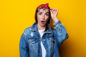 Surprised young woman in hair band, in denim jacket raised glasses on yellow background.