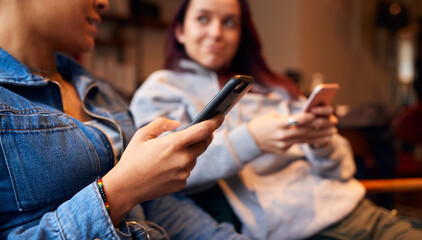 Close Up Of Same Sex Female Couple Sitting On Sofa At Home Looking At Mobile Phones Together
