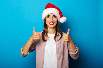 Welcoming young woman in shirt wearing santa claus hat showing thumbs up gesture on blue background.
