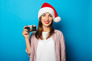 Smiling young woman in a shirt wearing a Santa Claus hat holding a retro camera on a blue...