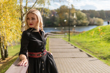 Fototapeta na wymiar A cheerful lady stands and leans on the railing, with a beautiful smile in black clothes, in the fall against the background of a pond with a blue sky.