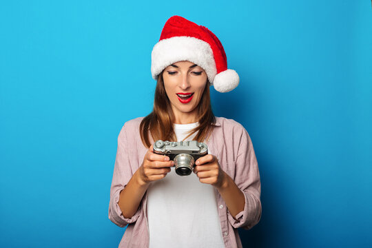 Young woman in santa claus hat looks in surprise at a retro camera on a blue background.
