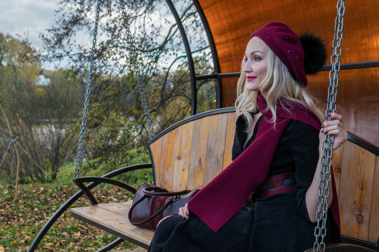 A beautiful lady sits on a garden swing, forged in a burgundy coat and biret