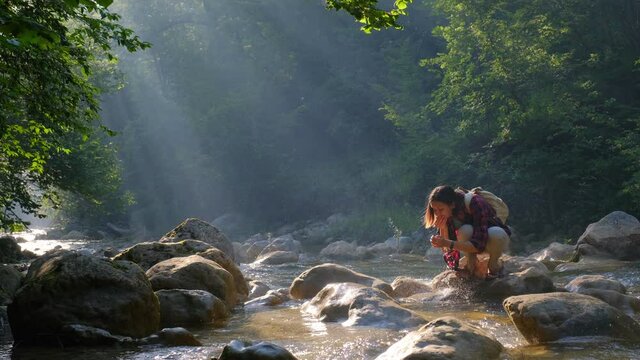 river or spring in summer forest. Female tourist with backpack drinking fresh mountain water while a hiking, scooped up cold transparent water with his hands. Concept of ecotourism travel