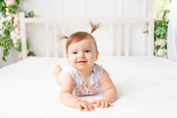 funny baby girl six months old lies in a bright beautiful room on a white bed in a lace bodysuit and smiles