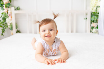 Fototapeta premium funny baby girl six months old lies in a bright beautiful room on a white bed in a lace bodysuit and smiles