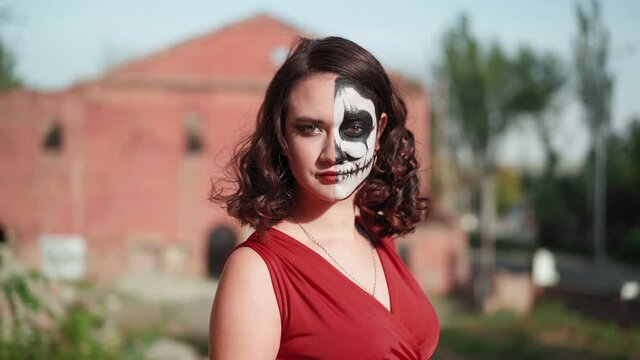Portrait beautiful slender woman in a red evening dress and awesome Halloween makeup stands against the backdrop of an abandoned building. Greasepaint for the Holiday on October 31. Slow motion