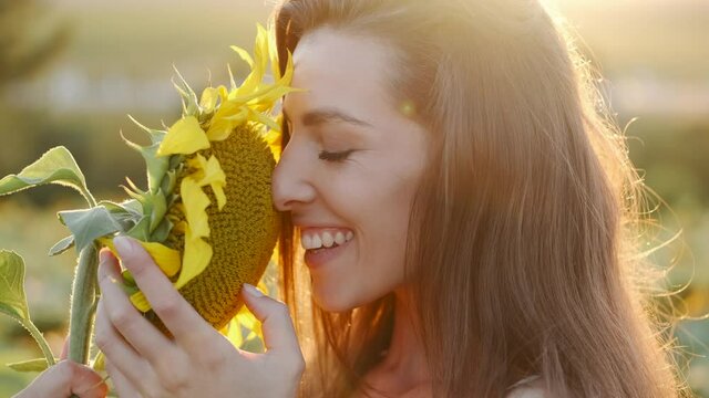 Beautiful young woman posing in field with sunflowers and enjoying nature. Woman smells flowers against background of nature and field of sunflowers. Portrait view. 4k.