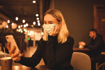 Young woman sits in a cafe, quarantine time. Happy blonde is resting in a cafe, wearing a mask and talking on the phone. Portrait photo of a woman in a black sweater
