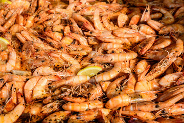 Close up of large portion of cooked shrimps with tomato sauce and yellow lemons in a large pan at a street food festival, ready to eat healthy seafood, beautiful orange monochrome outdoor background.
