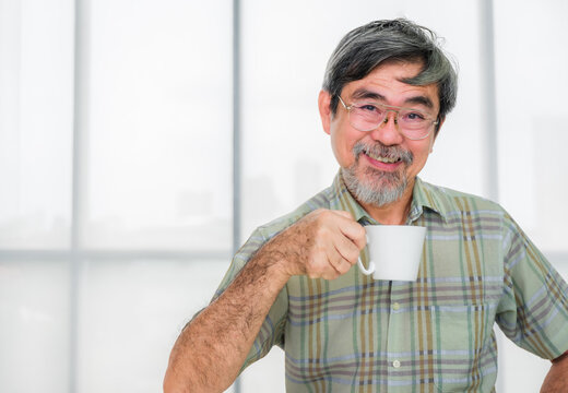Happy senior asian man sitting on couch and holding coffee cup ready to drink in living room. Elderly man gray hair beard and smile with coffee on sofa in morning. Lifestyle after retirement concept.