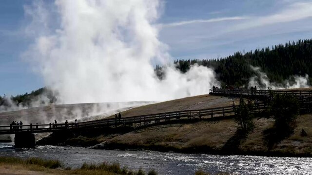 Tourists (unidentifiable) in silhouette cross river bridge to view steaming geothermal features under blue sky with high wispy clouds