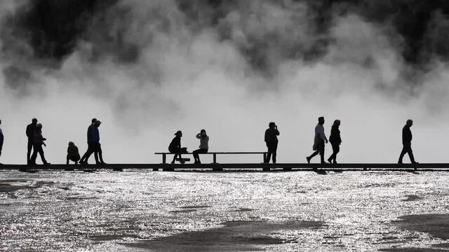 Tourists (unidentifiable) silhouetted between rising steam and reflecting water from hot springs walk boardwalk among geothermal features of the area