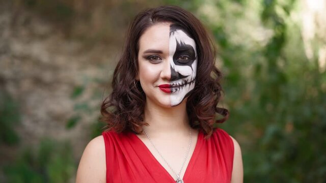Portrait beautiful woman in a red evening dress and awesome Halloween makeup stands and smiles against the backdrop of an abandoned building. Greasepaint for the Holiday on October 31. Slow motion