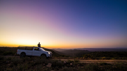 Fototapeta na wymiar Wild man sitting on the roof of a car during sunset in the mountains on an adventure