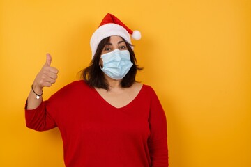 Beautiful middle aged woman wearing Christmas hat and medical mask standing against yellow...