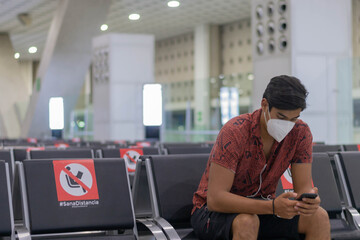 young man with face mask at the airport using the cell phone, with signs of healthy distance due to the pandemic