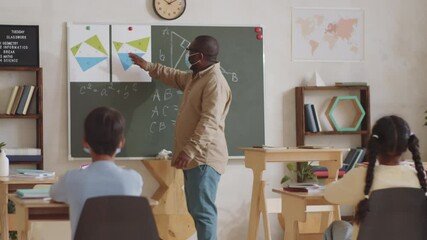 African American male teacher in medical face mask standing in front of children, pointing at chalkboard and explaining geometry lesson while working in school during coronavirus outbreak