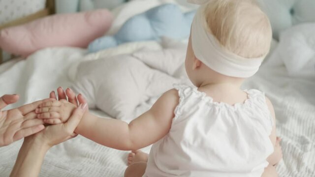 Close-up portrait of a small child in a light bodysuit from the back. Mom's gentle hands are holding the little hand of the baby. Mother and daughter together