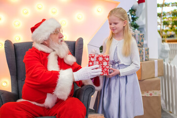 Fototapeta na wymiar Santa Claus gives girl a gift in Shopping Mall. Real authentic Santa talking and playing surprise games with kids . Christmas sales and wishes.