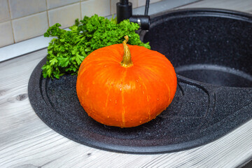 in the kitchen, near the sink, there is a ripe yellow pumpkin for cooking in the foreground and a bunch of green parsley in the background...