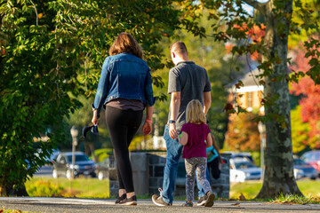 A young caucasian family of three is walking in the park at a lovely autumn afternoon. The cute little blonde girl follows her daddy and mommy. They all wear casual clothing.
