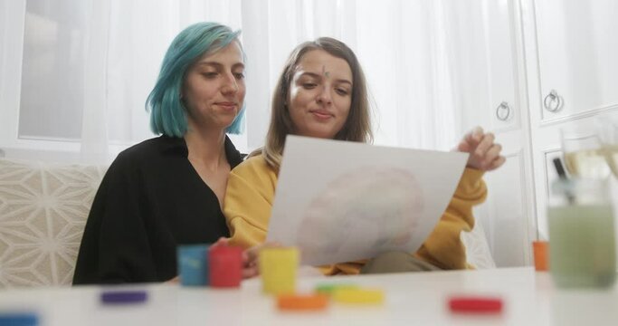 Two young lesbian women admiring an LGBT painting of the rainbow symbol which they have just painted together at a table in their living room at home before cuddling together in loving contentment