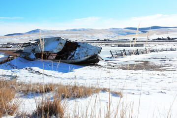 An old abandoned ship lying on the shore of Lake Baikal. Around the autumn steppe, village fences and snow-covered hills