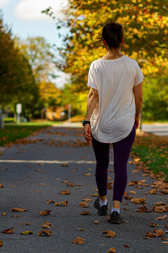 A young woman wearing sneakers, purple tight and a white blouse is walking alone on a hiking trail on a fall day. Path is covered with fallen leaves. A concept image for autumn exercise, walking alone